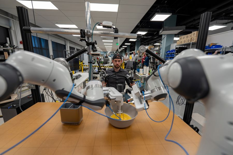 In a 2024 lab setting, a man intently operates a complex robotic system featuring multiple mechanical arms. He focuses on manipulating the AI-driven arms over a workspace with a bowl, while shelves stocked with tools reflect the latest AI adoption trends in robotics. | Ven Agency