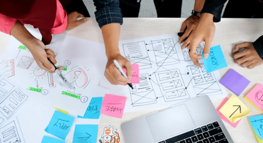 The Ultimate Guide to Website Layout and UX Design 1 A group of people collaborate around a table covered with design sketches, sticky notes, and a laptop, diving into the ultimate guide for perfect website layout. Hands point and write on the papers, indicating an active UX design brainstorming session. | Ven Agency