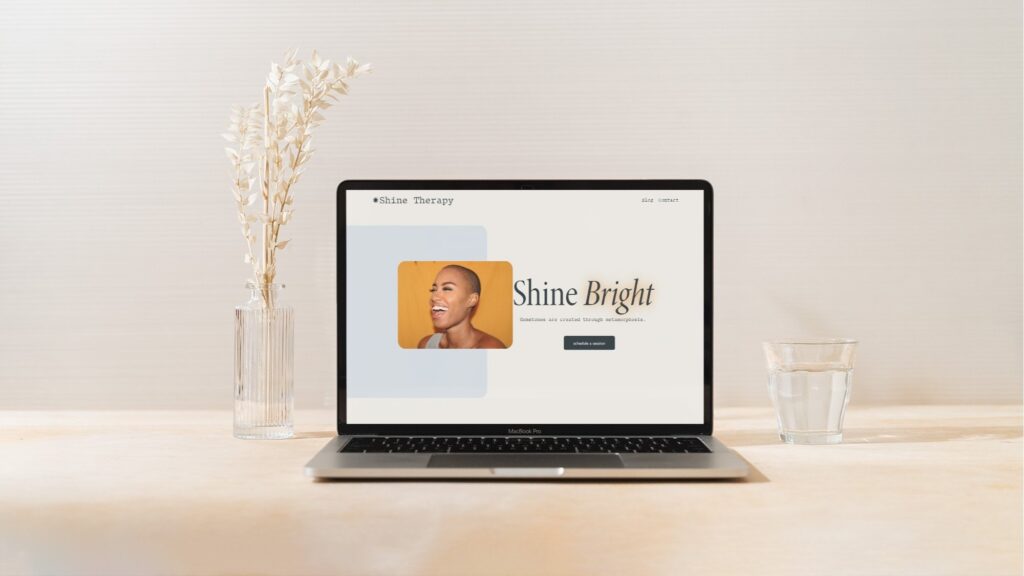 A laptop on a wooden table showcases a business website with the text "Shine Bright" beside an image of a smiling person. A tall vase with dried flowers sits to the left, and a glass of water to the right. This elegant setup, perhaps crafted on Squarespace, is framed by a neutral-toned background. | Ven Agency
