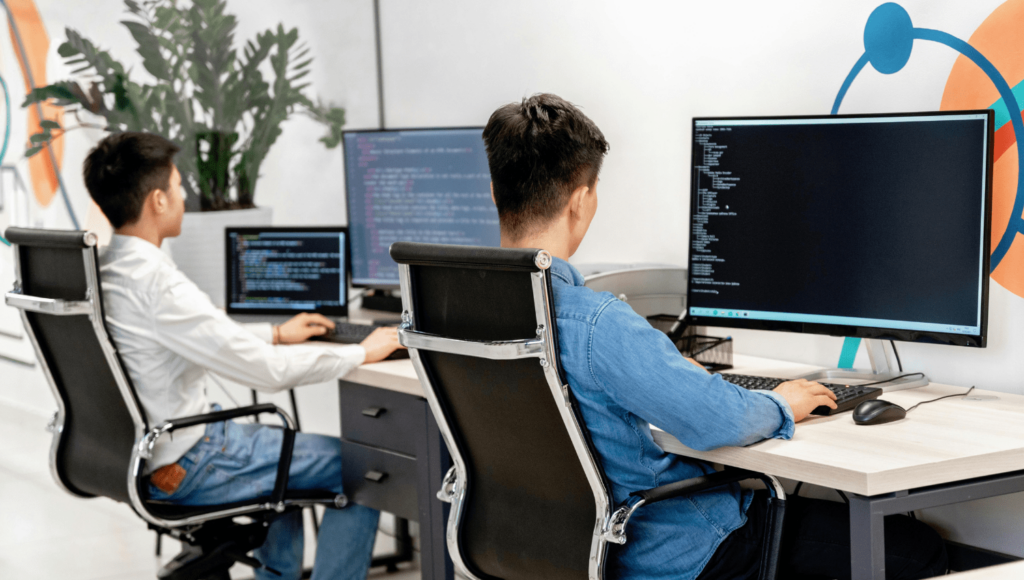 Two people are seated at desks working on desktop computers, crafting a business website on Squarespace. Both screens display lines of code. There's a large plant in the background and colorful wall art, giving the modern tech workspace an inspiring atmosphere. | Ven Agency