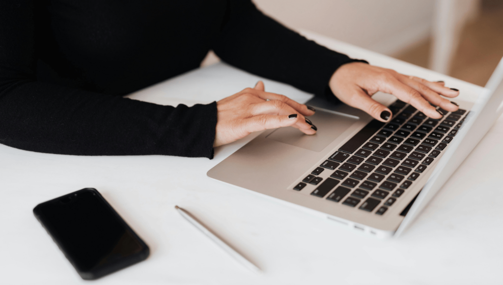 A person types on a laptop, crafting their business website with both hands on the keyboard. The laptop sits on a white desk, alongside a smartphone and pen, as they wear a black long-sleeve shirt. Squarespace tools lay at their fingertips. | Ven Agency